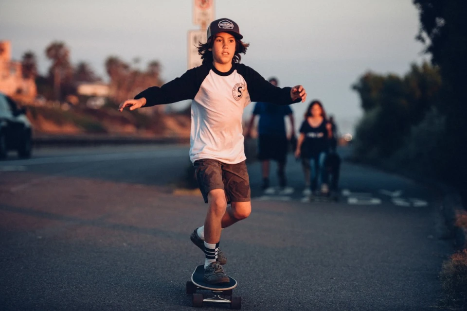 A boy skateboarding on a road with people walking in the background during sunset, wearing a baseball cap and casual clothing.