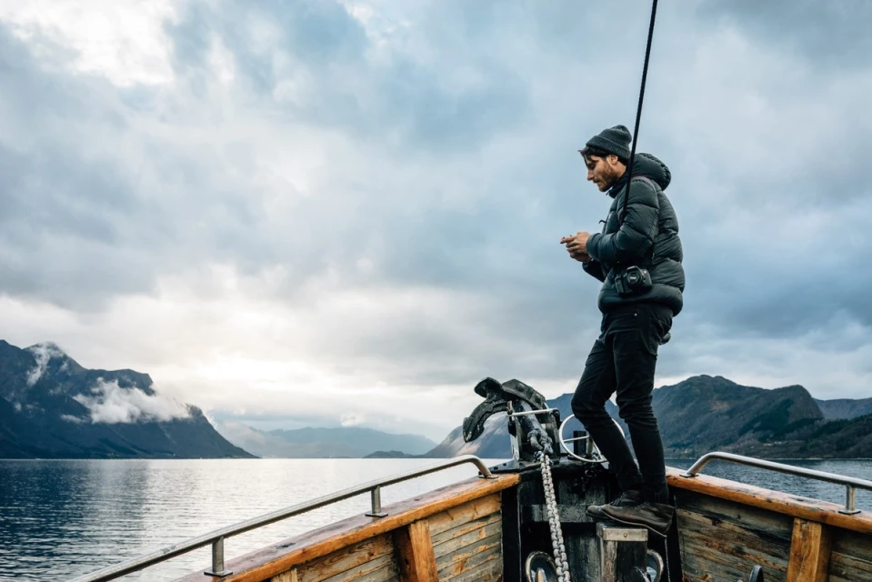 Man standing on a boat deck with mountains and cloudy sky in the background, wearing a puffer jacket and beanie, looking at a cell phone.