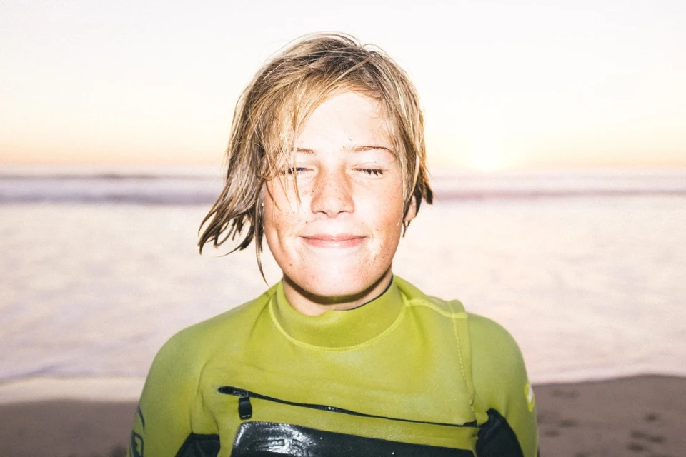 Close-up of a surfer in a wetsuit standing on a sandy beach with closed eyes, featuring a soft sunset background and serene atmosphere.
