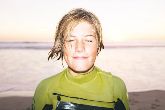 Close-up of a surfer in a wetsuit standing on a sandy beach with closed eyes, featuring a soft sunset background and serene atmosphere.