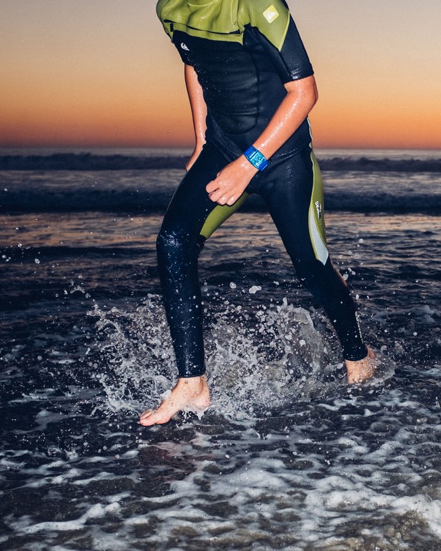 Teenage surfer wearing a wetsuit walks in shallow ocean waves with a setting sun creating a beautiful orange color in the sky.