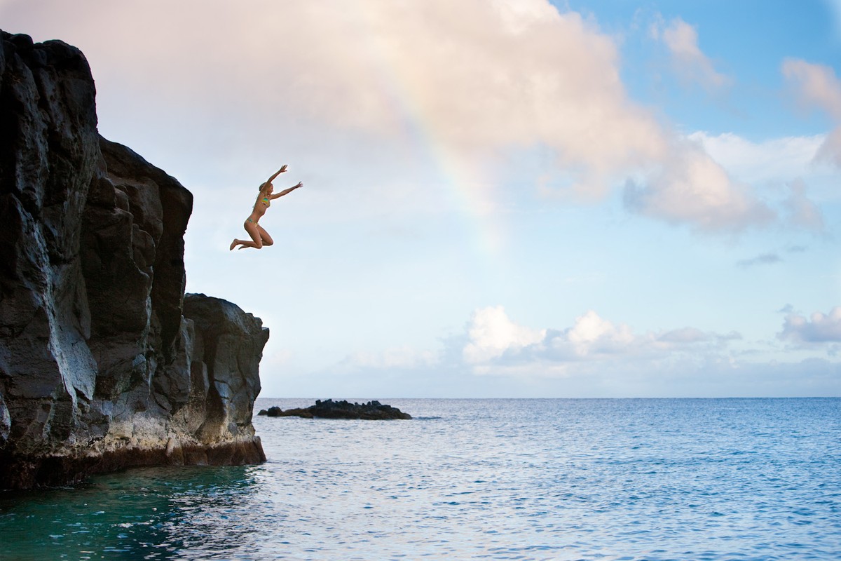 Woman jumps from a cliff into the ocean on a sunny day with a blue sky and a partial rainbow, capturing a moment of outdoor adventure.