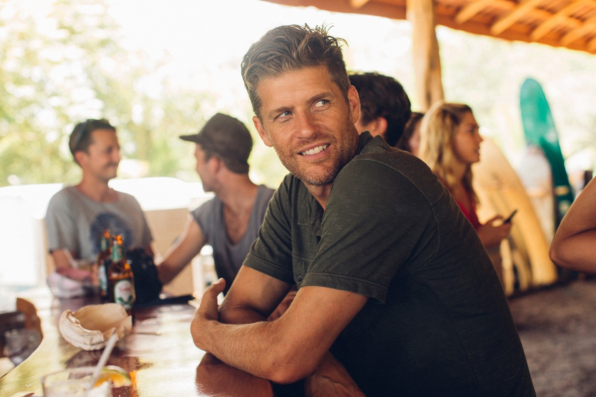 Portrait of smiling man with friends in outdoor cafe setting; casual clothing, tropical vibes, and social gathering at wooden table.
