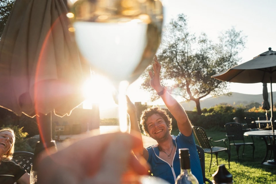 A man raises a wine glass in celebration outdoors, with a bright sunny backdrop of trees and rolling hills; drinks are on the table.