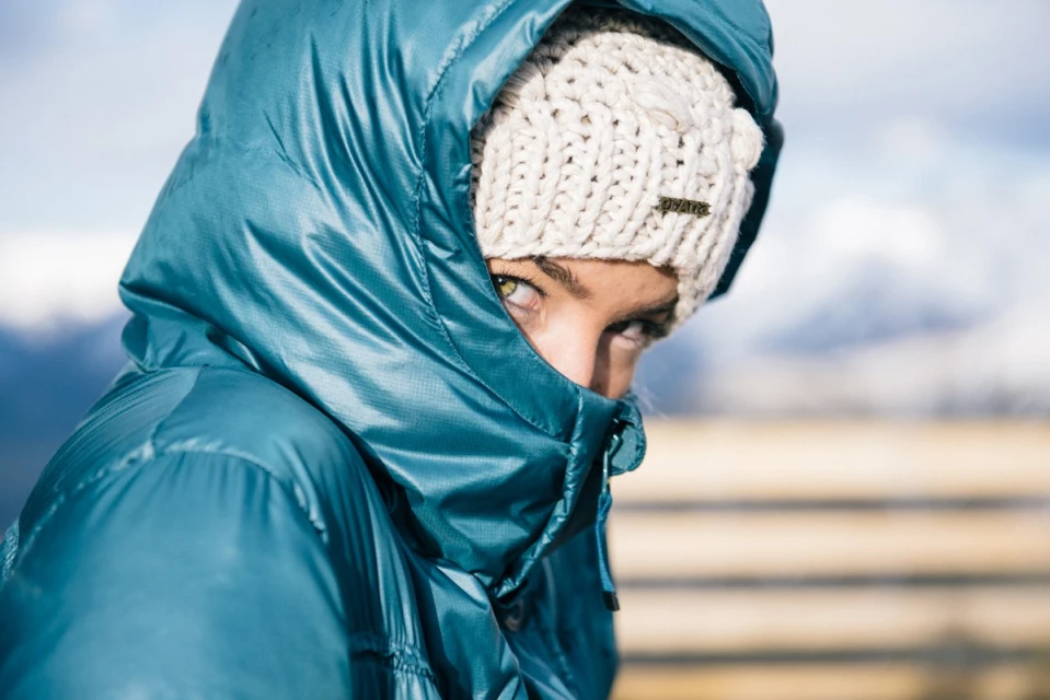 Close-up shot of a woman in a hooded teal puffer jacket and a cream-colored knit beanie, partially obscuring her face with focus on her eyes.