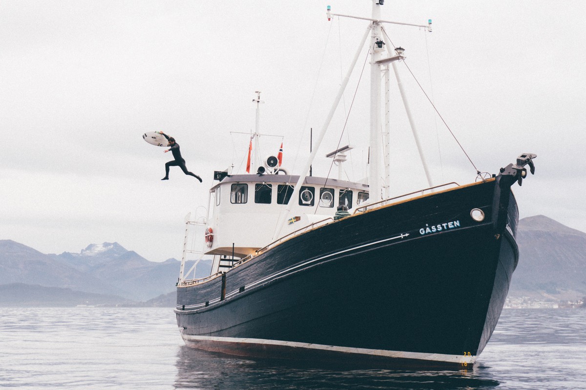 Man in wetsuit jumps with surfboard from boat into water, mountains in the background under a cloudy sky, in a wide shot.