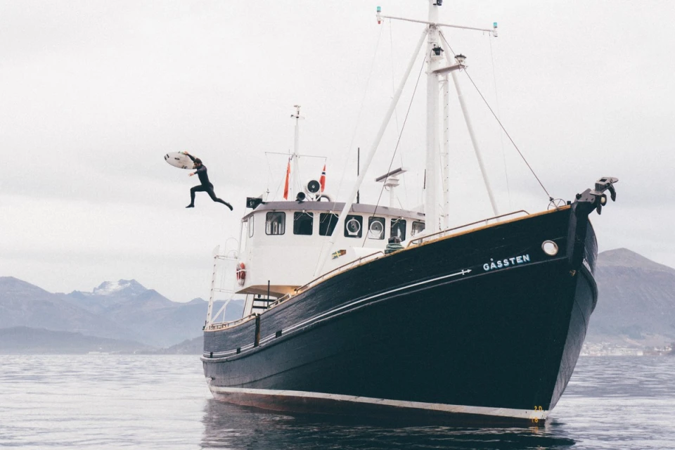 Man in wetsuit jumps with surfboard from boat into water, mountains in the background under a cloudy sky, in a wide shot.
