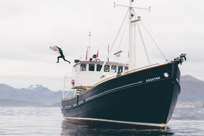 Man in wetsuit jumps with surfboard from boat into water, mountains in the background under a cloudy sky, in a wide shot.