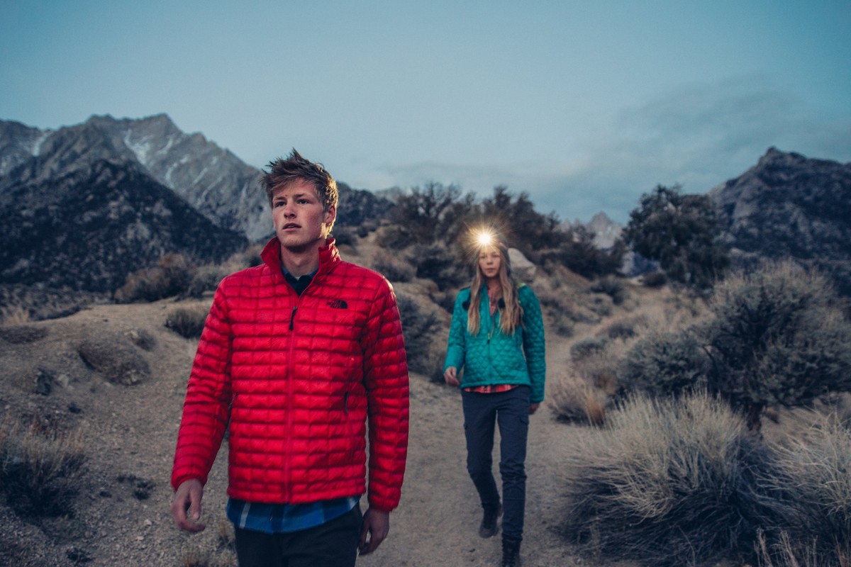 Two hikers, a man and woman, traverse a rugged trail at dusk, wearing outdoor jackets and headlamps, set against a backdrop of mountains.