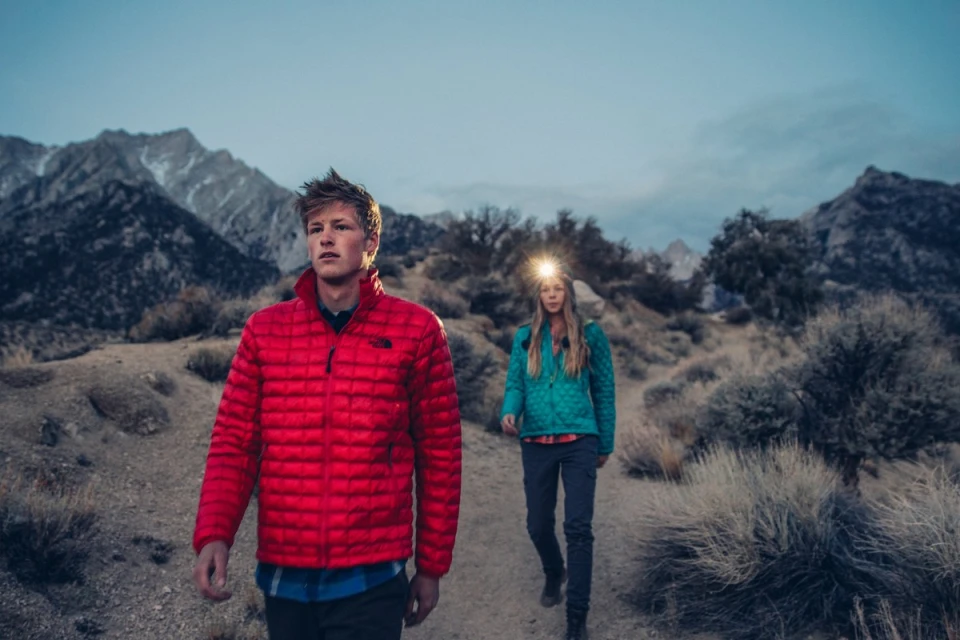 Two hikers, a man and woman, traverse a rugged trail at dusk, wearing outdoor jackets and headlamps, set against a backdrop of mountains.
