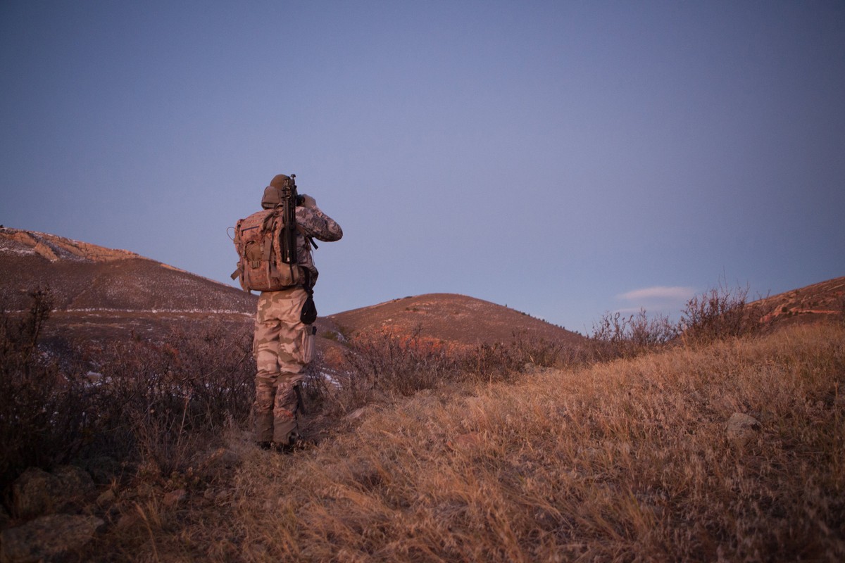 Man in camouflage stands on a grassy hilltop at dusk, facing away from the camera, wearing a backpack, with mountains in the distance.