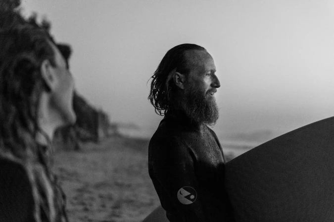 Black and white shot of two surfers on a beach at dusk; one man in wetsuit, looking out to the ocean and holding surfboard.