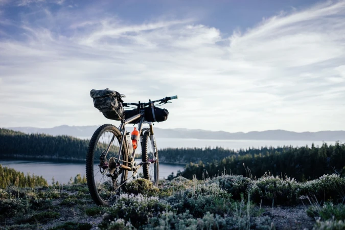 Bike packed touring bicycle rests at scenic overlook with view of lake and forest against cloudy sky in outdoor environment.