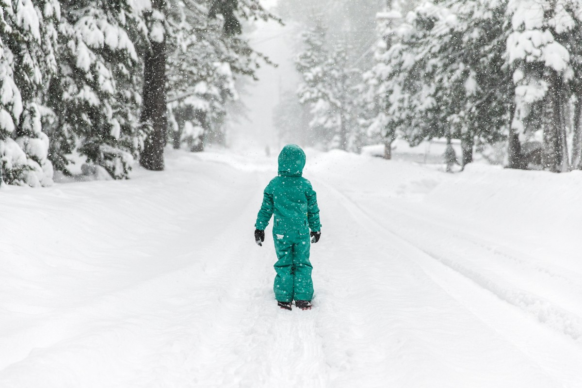 Child in teal snowsuit walks down snowy path surrounded by snow-covered trees in a winter forest during a heavy snowfall.