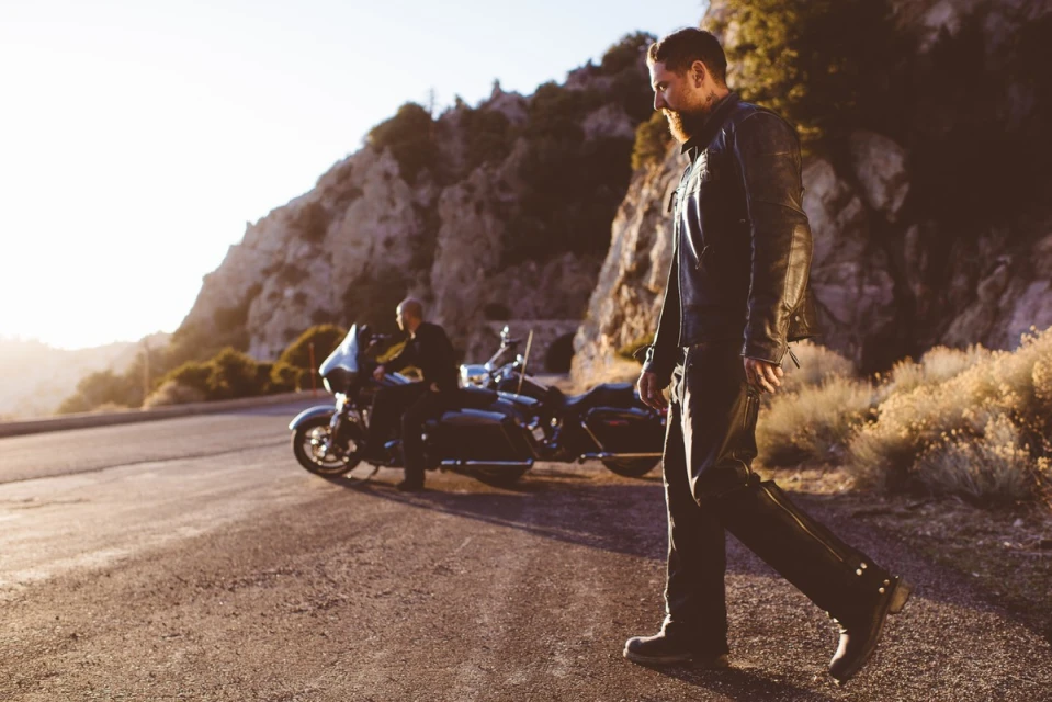 An adult male walks along a road next to a parked motorcycle while another adult male sits on the bike near a mountainous landscape.
