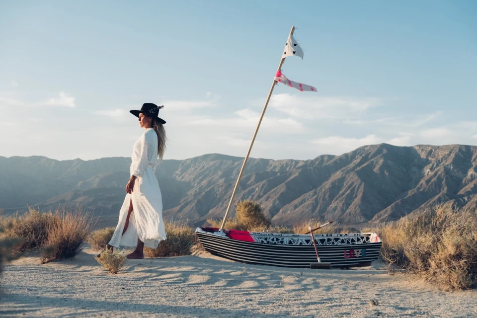 A woman in a white dress and hat stands next to a boat on a sandy surface with mountains in the background on a sunny day.