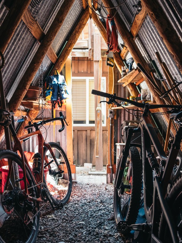 A-frame shed interior featuring two bicycles, tools, storage, and wood construction, with light filtering through a window in the background.