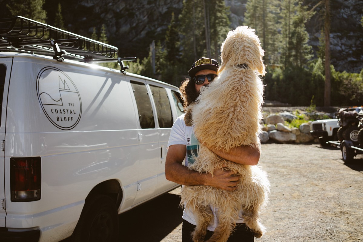 Man holding a fluffy dog in front of a white van near a forest. The van has a logo for The Coastal Bluff on its side.
