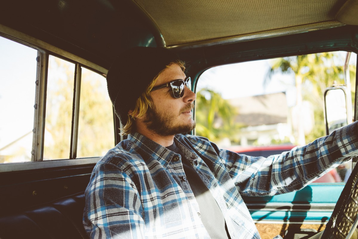 Man in black beanie and sunglasses drives vintage truck, wearing plaid shirt. He steers with the steering wheel on a sunny day.
