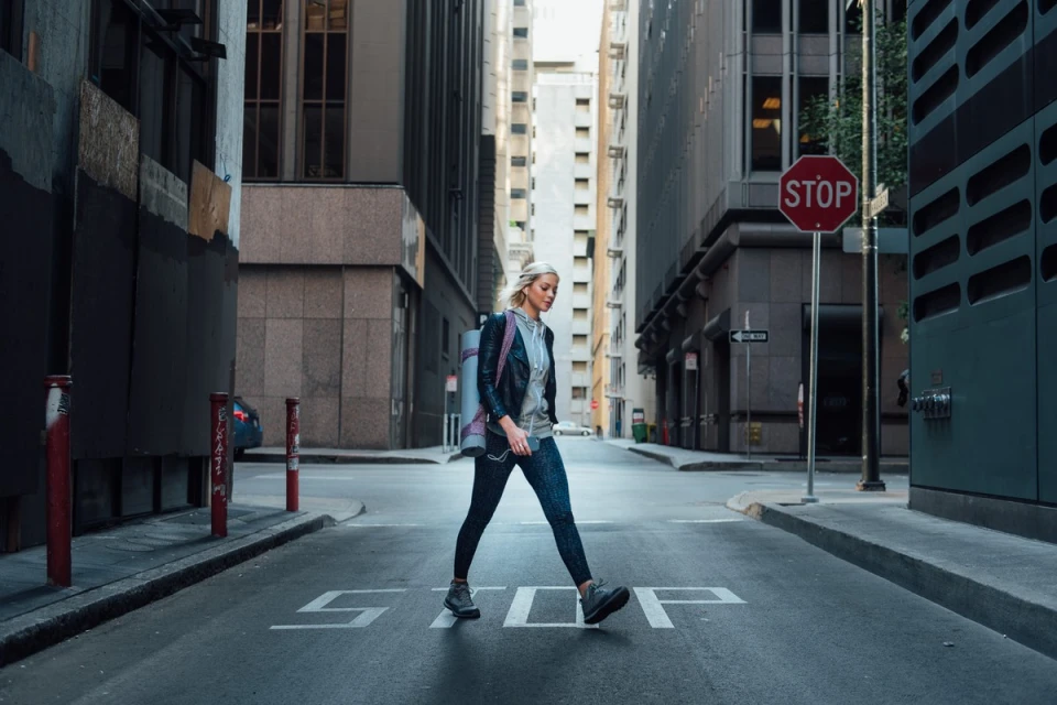 Fit young woman wearing workout clothes and carrying a yoga mat walks across a city street in front of urban buildings.