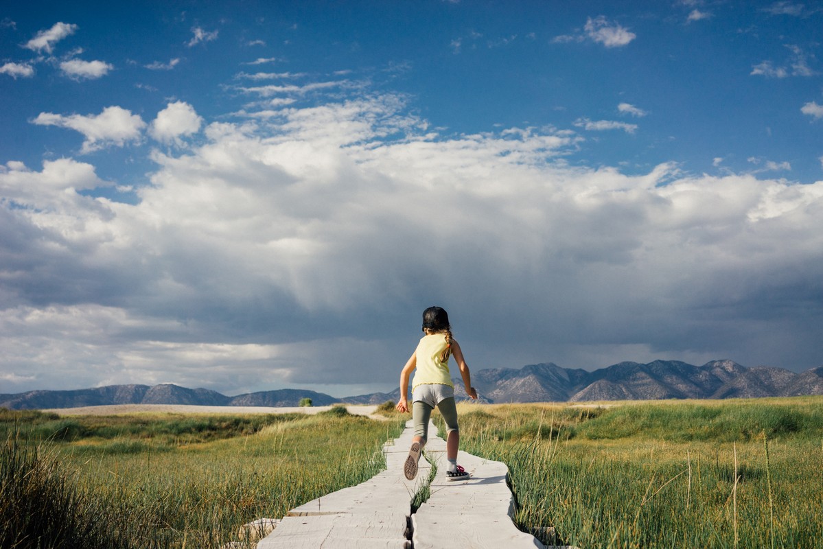 A child runs along a wooden walkway surrounded by a field of tall grass under a cloudy blue sky, with mountains in the distance.