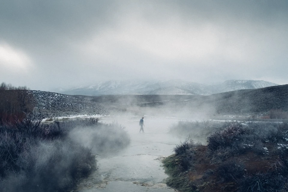 Person walks through steaming geothermal landscape with low clouds and snow-dusted hills in the background on an overcast day.