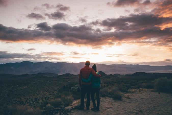 A man and woman standing together watching sunset over a mountain range; the couple is wearing casual clothing.