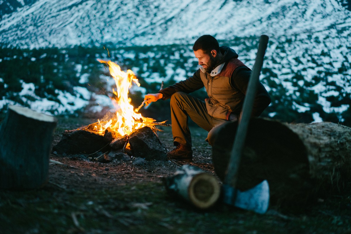 Man in vest crouches by a bright campfire at dusk with an axe and logs nearby, with snow-covered mountains in the background.