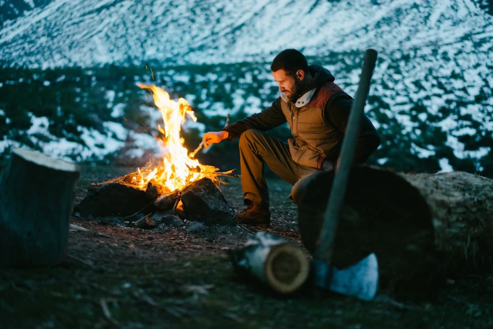 Man in vest crouches by a bright campfire at dusk with an axe and logs nearby, with snow-covered mountains in the background.