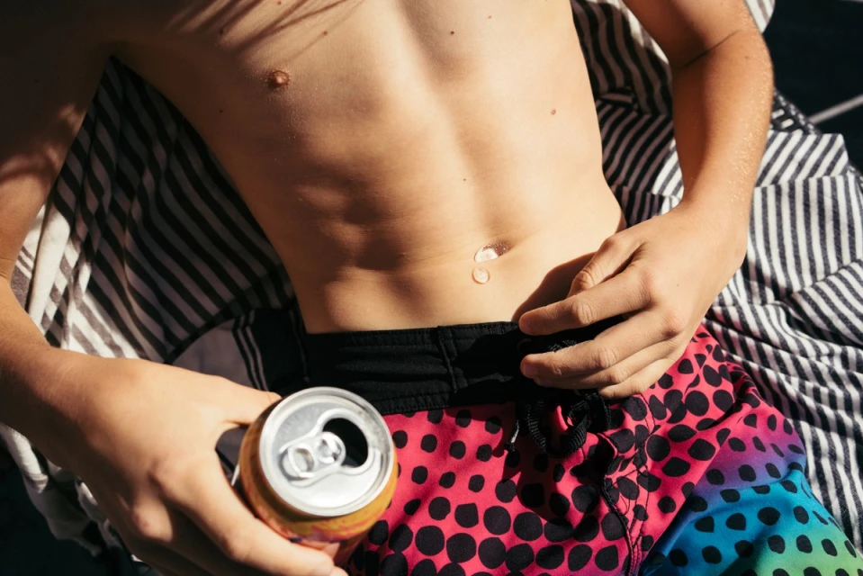 Young man in swim trunks lying on a striped towel, holding a can of soda on a sunny day with strong shadows from overhead foliage.