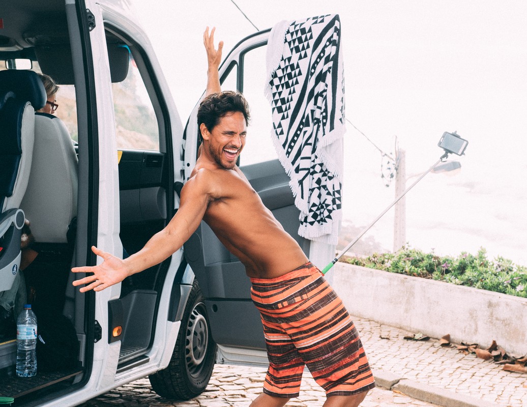Enthusiastic man poses by white van with towel and selfie stick, near the beach, exhibiting joyful expression and casual beach attire.