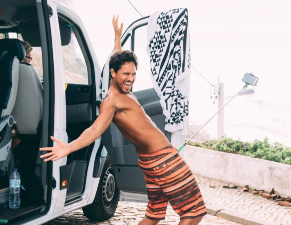 Enthusiastic man poses by white van with towel and selfie stick, near the beach, exhibiting joyful expression and casual beach attire.