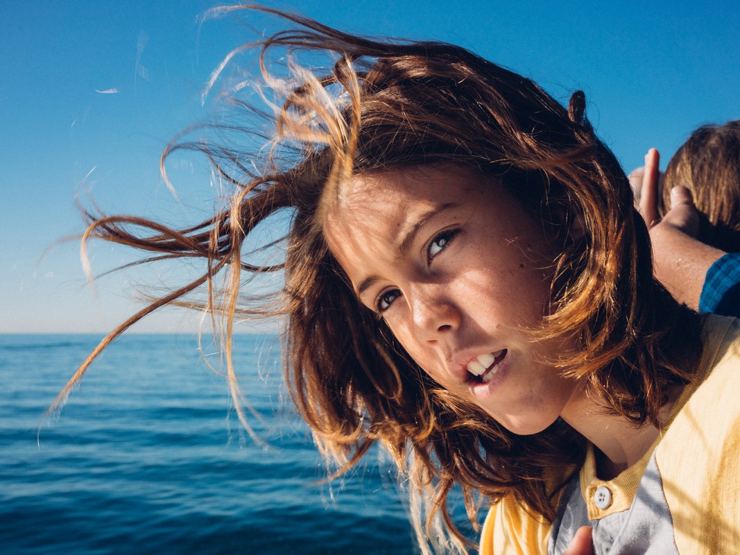 Close-up shot of a young child with windblown brown hair on a boat, with blue sky and ocean in background. Child looks directly at the camera.