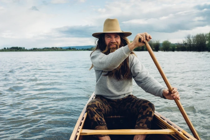 A person wearing a hat paddles a canoe on a lake with forested shoreline under a cloudy sky, showcasing an outdoor water sport activity.