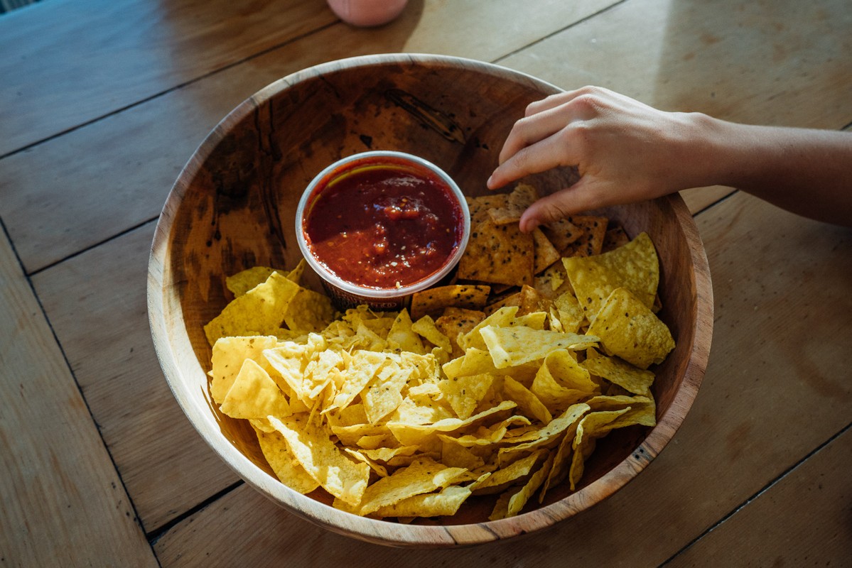 Overhead shot of wooden bowl with tortilla chips and a container of red salsa, with a hand reaching in to grab a chip.