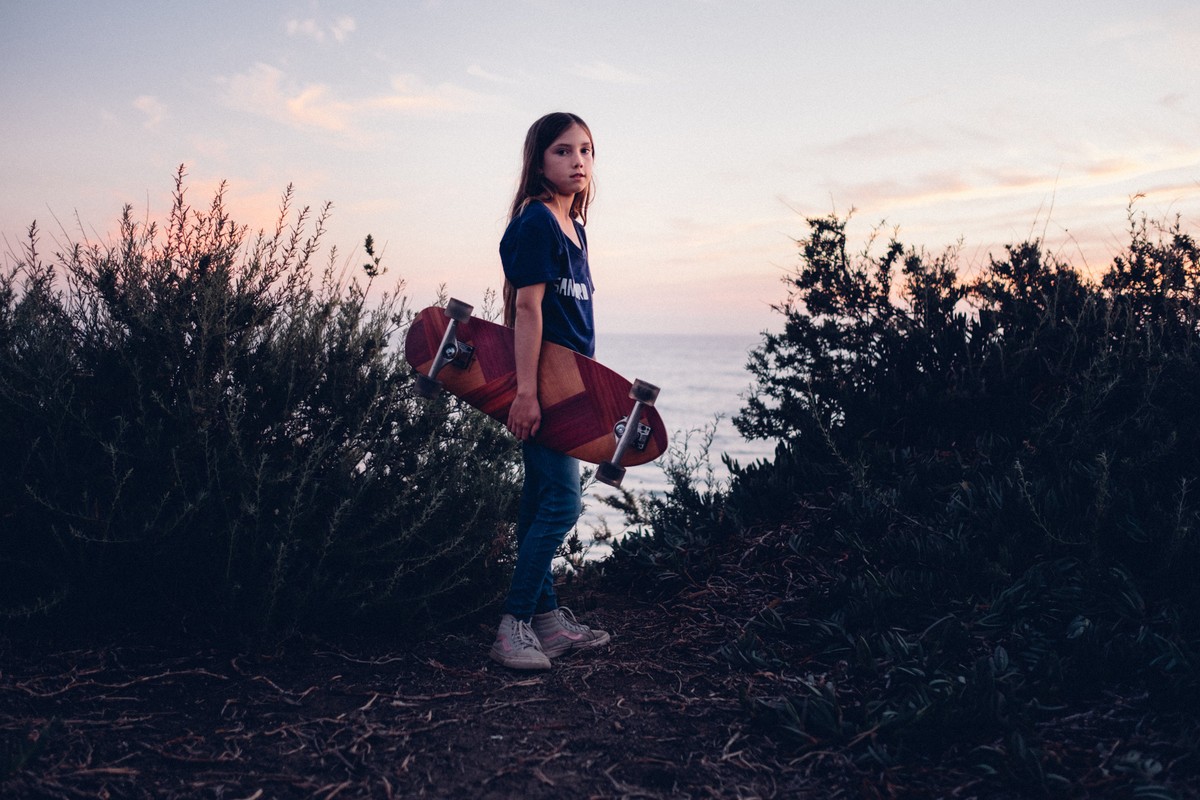 Young girl stands with a skateboard on a path near a grassy overlook above the ocean at dusk with a pink and orange sky.
