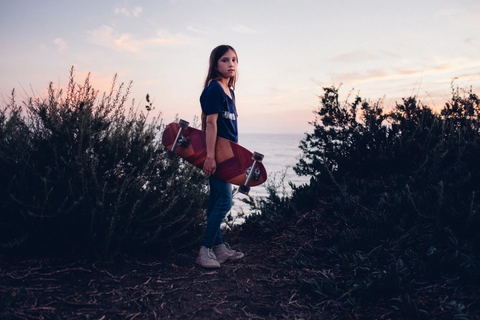 Young girl stands with a skateboard on a path near a grassy overlook above the ocean at dusk with a pink and orange sky.
