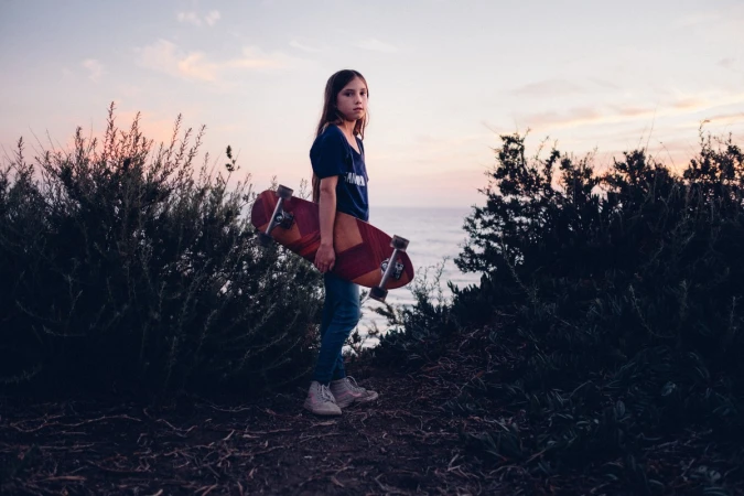 Young girl stands with a skateboard on a path near a grassy overlook above the ocean at dusk with a pink and orange sky.