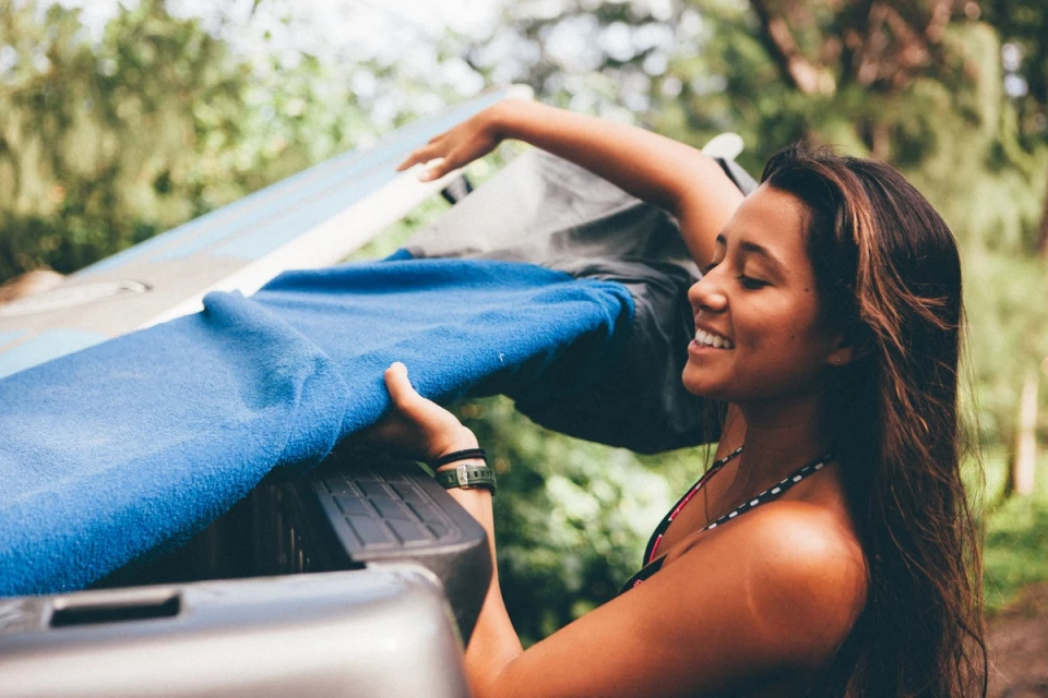 Smiling woman removing a blue blanket from the back of a pickup truck outdoors in a green, wooded area on a sunny day.