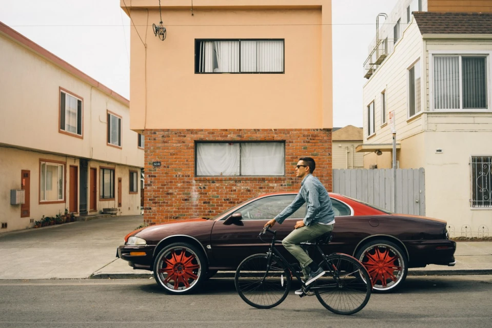 An adult male rides a bicycle on a city street near a parked maroon car and buildings in the background on an overcast day.