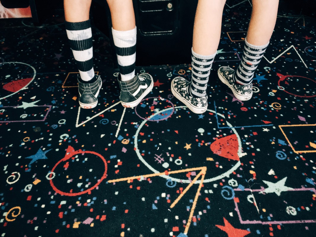 Close up on two pairs of legs wearing striped socks and sneakers standing on a carpet with colorful geometric shapes and stars.