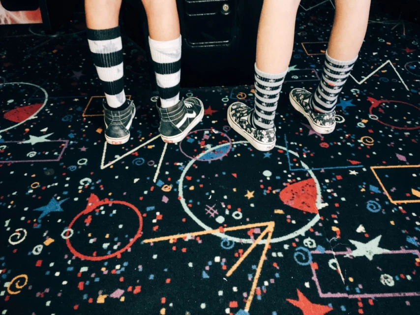 Close up on two pairs of legs wearing striped socks and sneakers standing on a carpet with colorful geometric shapes and stars.