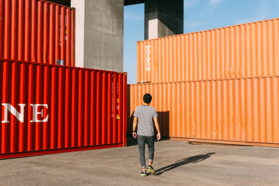 Man walks toward stacked red and orange shipping containers under a concrete structure against a clear blue sky on a sunny day.