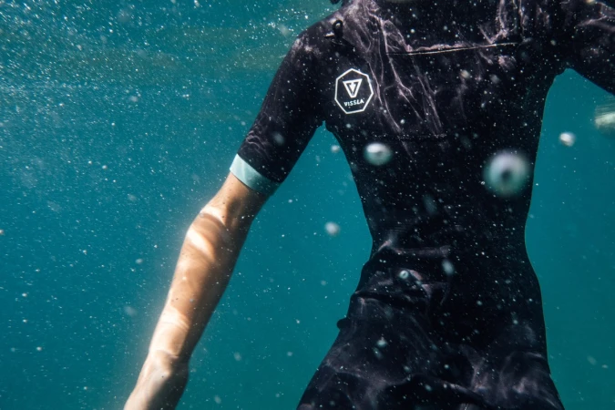 Person in a black wetsuit swimming underwater in clear turquoise water with visible bubbles; Vissla logo is visible on the suit.
