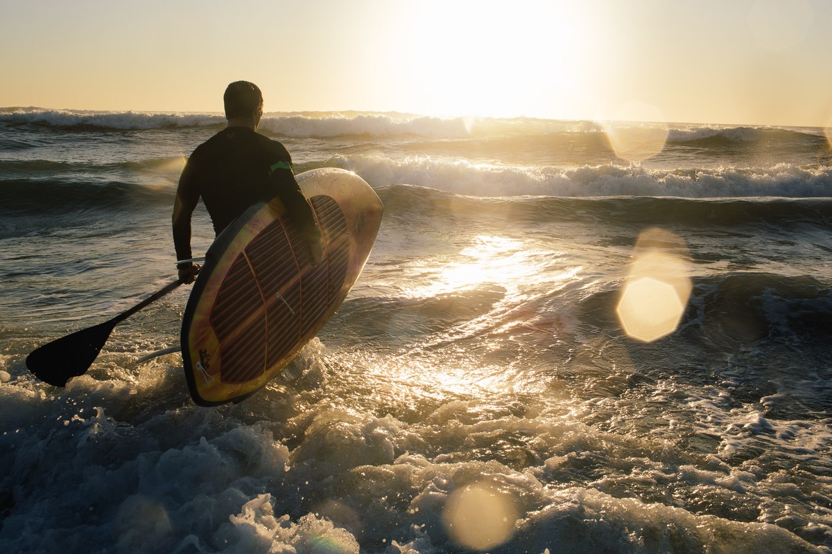 Adult male in wetsuit carries paddleboard and paddle into ocean waves at sunset, creating golden backlight and water reflections.
