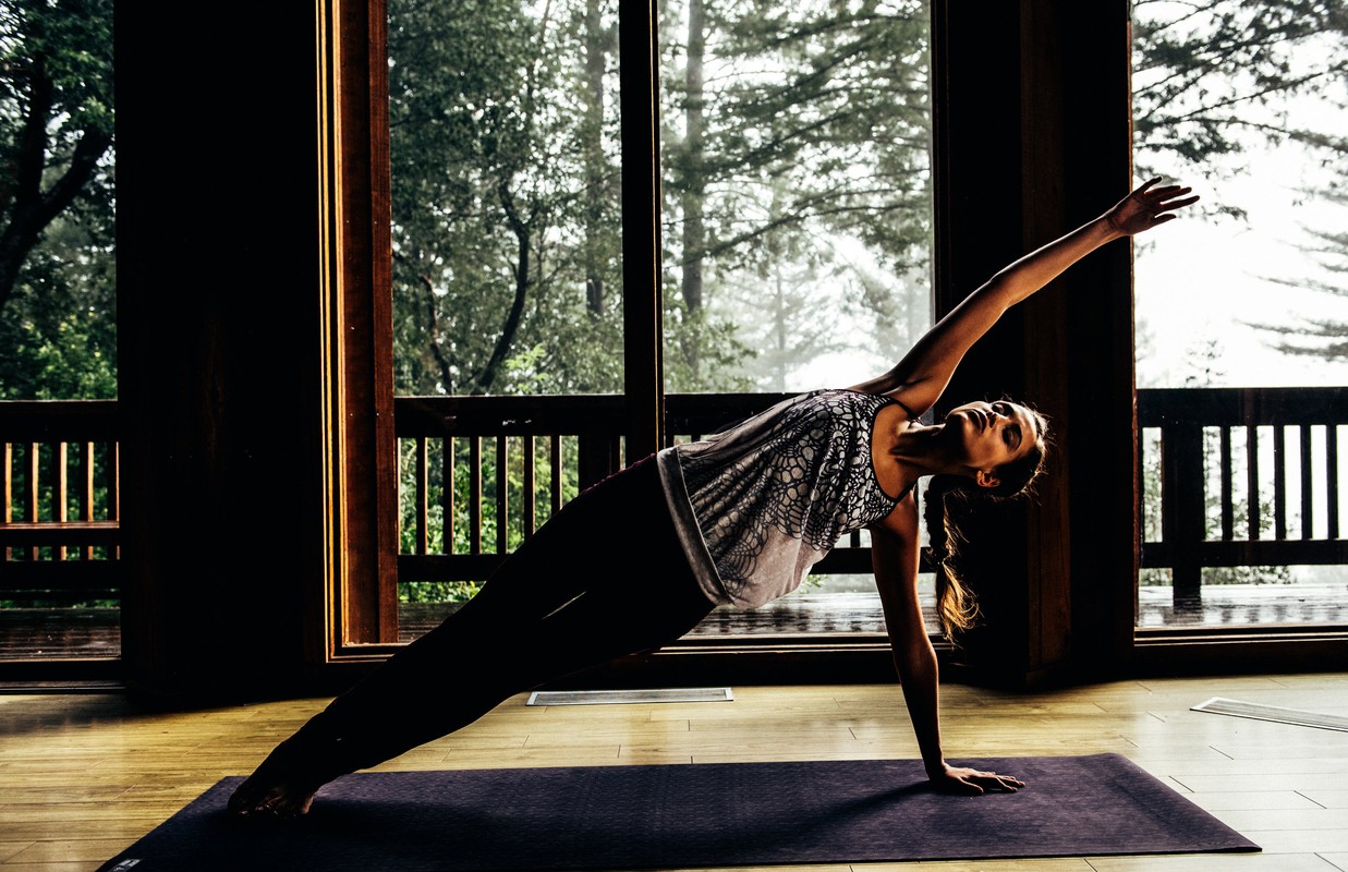 Adult woman performing side plank yoga pose on a mat indoors with a windowed backdrop offering a view of a forest. 
