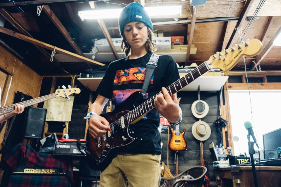 Teenager playing electric guitar in music space, captured in a medium shot with natural lighting and other music equipment visible.