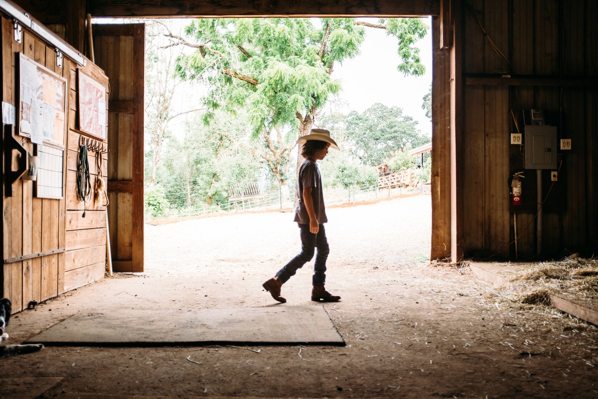 Young child in a cowboy hat walks through an open barn doorway, toward trees and sky in the background on a sunny day.