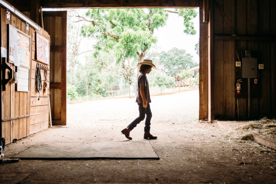Young child in a cowboy hat walks through an open barn doorway, toward trees and sky in the background on a sunny day.