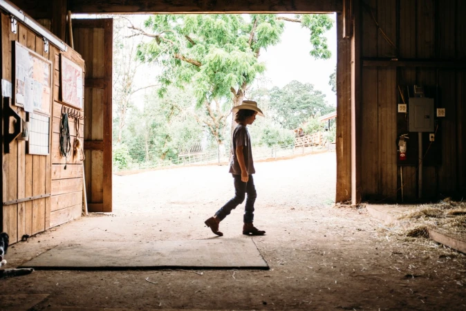 Young child in a cowboy hat walks through an open barn doorway, toward trees and sky in the background on a sunny day.